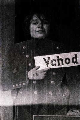 Black-and-white portrait of an elderly woman holding a handmade sign reading "Vchod" (Czech for "Entrance"). She wears a patt...