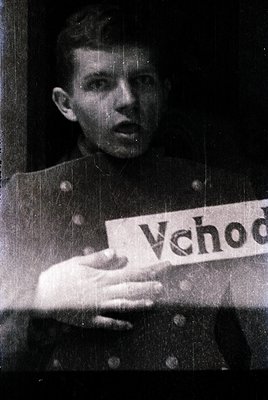 Young man in mid-20th-century military uniform holds a handmade sign reading "Vchod" (Czech/Slovak for "Entrance"). Uniform s...