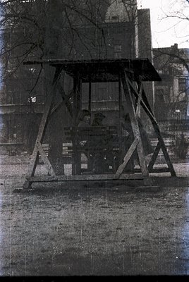 Vintage outdoor swing set with metal frame and wooden slats, likely mid-20th century. Surrounded by bare trees and a multi-st...