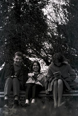 Vintage black-and-white photo of three individuals seated on a wooden bench beneath leafless trees, suggesting late autumn/wi...