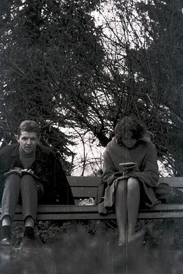 Two individuals seated on a wooden bench in a park-like setting, mid-20th century. The man wears a dark coat, trousers, and a...