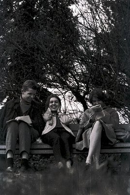 Three individuals seated on a stone bench in a park, mid-20th century. Left: man in military-style jacket, reading. Center: w...