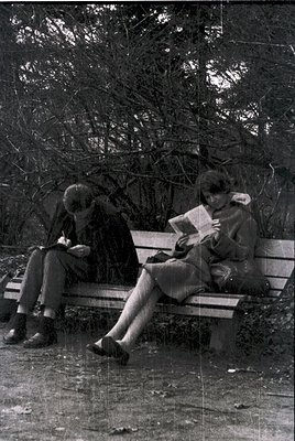 Two individuals reading outdoors on a wooden bench in a park setting, likely mid-20th century. The vintage sepia tone suggest...