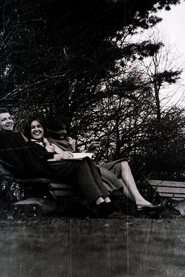 Three individuals pose casually on a park bench in winter, surrounded by bare trees. The woman in the center wears a dark bla...