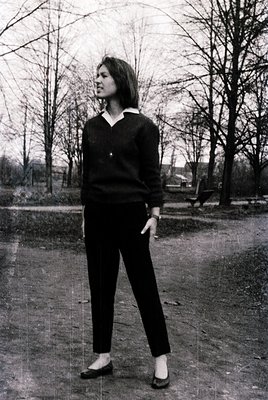 Portrait of a woman in a V-neck sweater and tailored trousers, standing outdoors in a park-like setting with leafless trees. ...