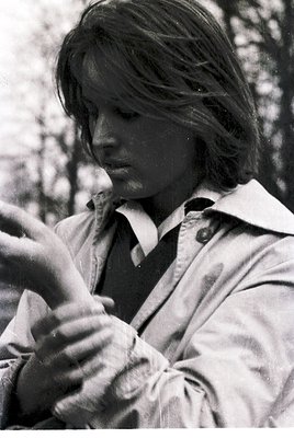 Young individual in mid-1970s outdoor setting, adjusting a camera strap over shoulder. Blurred foliage background suggests wo...