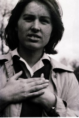 Portrait of a person in a vintage jacket, hands clasped over chest, mid-speech. Black-and-white, likely 1960s–1970s. Soft foc...