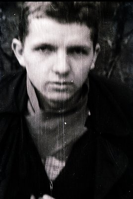 Black-and-white portrait of a young man in a dark, high-collared coat, likely from the 1940s–1950s. His expression is neutral...