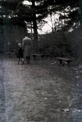 Vintage black-and-white photo of an adult and child walking on a paved path in a wooded park. The adult wears a light-colored...