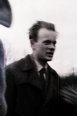 Vintage black-and-white portrait of a young man in a textured blazer, mid-speech outdoors. Blurred background suggests motion...