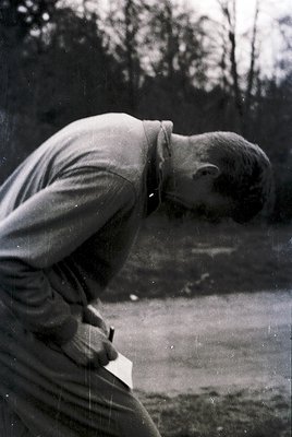 Mid-20th century man in outdoor setting, bending to adjust a large, flat object (likely a sign or board) with a pencil. His a...
