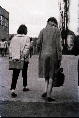 Two individuals walk away from the camera in a mid-20th-century urban setting, likely the 1960s–70s. The woman wears a knee-l...