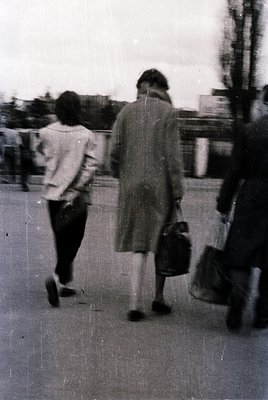 Blurred mid-century street scene featuring two women in motion—one in a light blouse and dark skirt, the other in a long coat...