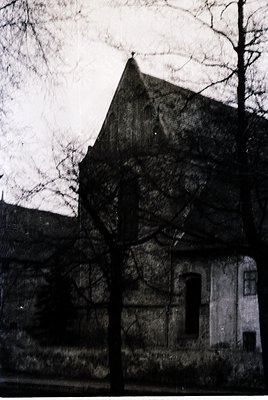 Black-and-white shot of a weathered, gabled building with exposed brickwork and arched windows, likely mid-20th century. Leaf...