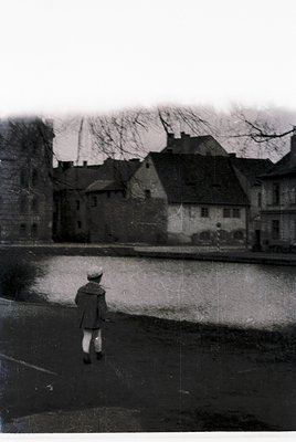 Vintage black-and-white street scene featuring a lone child walking away from camera, dressed in mid-20th-century attire (hat...