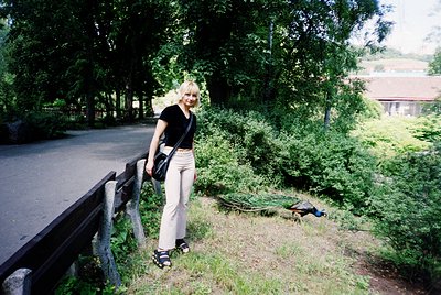 Woman in 1990s-style light-wash trousers and black top poses by a rustic wooden railing beside a paved path. Lush greenery an...