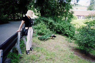Woman in 1990s-style casual attire (black top, high-waisted beige pants, sandals) leans on a metal railing, observing a small...