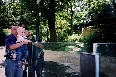 Family group at a zoo enclosure, likely a reptile exhibit, with a man holding a baby and another child standing beside him. T...
