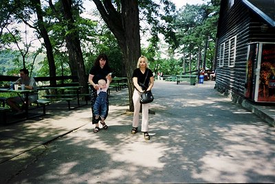Two women stroll along a shaded park pathway lined with wooden benches and tall trees. The woman on the left holds a child in...