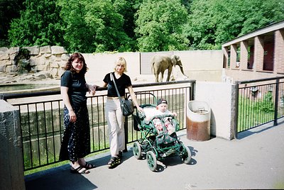 Two women pose with a child in a stroller at a zoo enclosure, likely late 1990s–early 2000s. The adult on the left wears a bl...