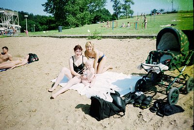Two women pose on a sandy beach in 1990s-era swimwear, seated on a white towel. One wears a black bikini, the other a white o...