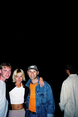 Four individuals pose against a dark background, likely an outdoor night event. The central male wears a denim jacket over an...