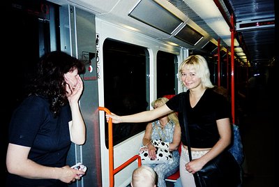 Vintage metro interior with two women laughing near an open door; one holds a small child. Red poles, white seats, and a clas...