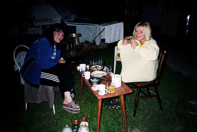 Two individuals enjoying an evening meal outdoors under a tented canopy. The woman on the left wears a blue Adidas tracksuit ...