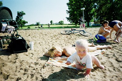 Sunlit beach scene with two adults and a toddler on a sandy area. Adults lie on towels, one reading a book, while the toddler...