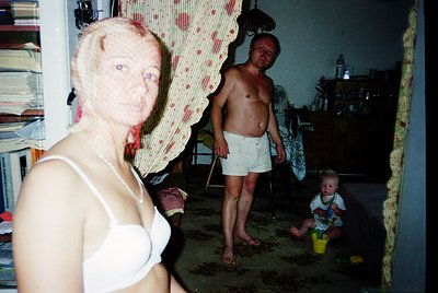 Intimate family moment captured in a modest indoor setting, likely mid-20th century. A woman in a white bra stands beside a m...