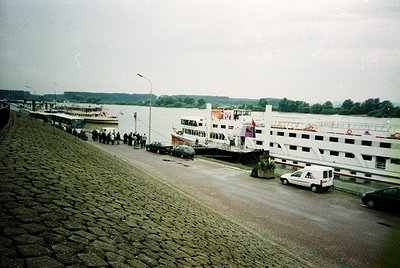 Riverfront passenger ferry terminal with cobblestone quay, featuring mid-20th century vessels docked alongside. Crowds gather...