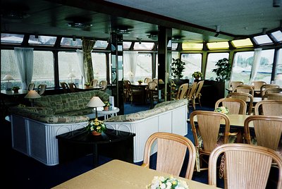 Luxurious mid-20th century river cruise lounge with panoramic windows showcasing water views. White paneling, wicker chairs, ...