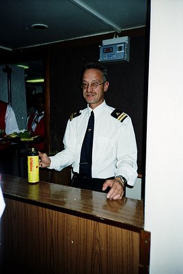 Maritime officer in white uniform with black stripes and epaulettes stands behind a wooden counter aboard a ship, holding a y...