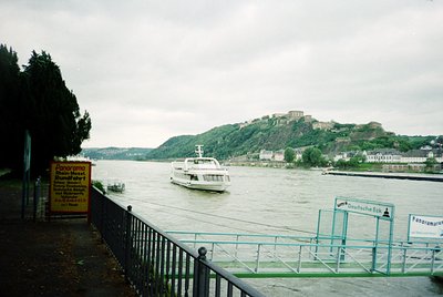 Riverfront ferry crossing near a hilltop fortress, flanked by historic European architecture. Prominent "Deutsche Eck" signag...