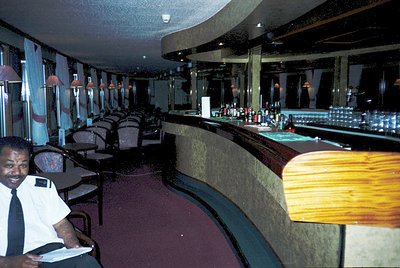Mid-century cruise ship lounge with curved wooden bar and pendant lighting. Staff in uniform (white shirt, black apron) stand...