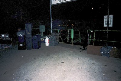 Empty nighttime ferry terminal with stacked luggage and cargo. Overhead signage reads "Tel. 0211" and "FERRY". Barriers and r...