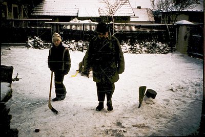 Two individuals navigate a snow-covered residential yard, likely mid-20th century. The adult wears a heavy winter coat, hat, ...