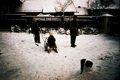 Vintage winter scene: three individuals in winter attire clearing snow from a residential courtyard. Snow-covered wooden fenc...