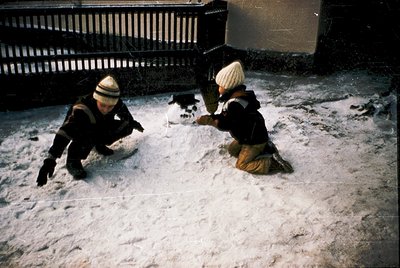 Two children build a snowman in an urban courtyard, surrounded by snow-covered pavement and a wrought-iron fence. Mid-20th ce...