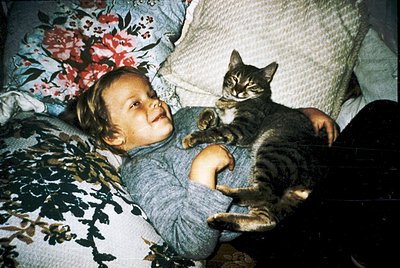 Vintage candid of a child and tabby cat sharing a cozy indoor moment on floral-patterned bedding. Warm, nostalgic lighting su...