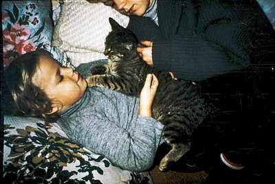 Vintage indoor scene featuring a child and kitten in close contact on a floral-patterned bed. The child appears relaxed, hold...