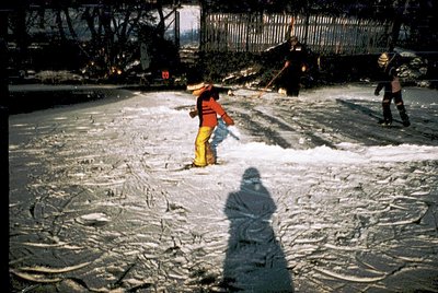 Three workers clear snow from a paved urban surface using shovels, likely in a 1960s–1970s city. Bright winter gear—yellow pa...