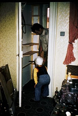 Mid-20th century kitchen interior with two adults: man in khaki shirt adjusting shelving, woman in polka-dot apron and white ...