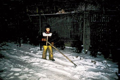 Industrial worker in yellow snow gear shoveling snow near a rusted metal fence and dilapidated brick structure. Snow-covered ...