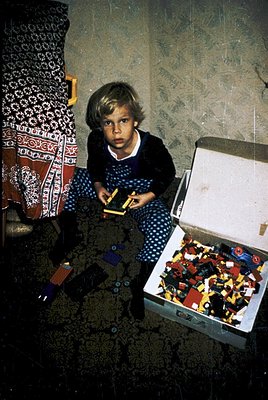 Child in mid-20th-century indoor setting, seated on patterned shag carpet, holding a wooden toy train. Open cardboard box bes...