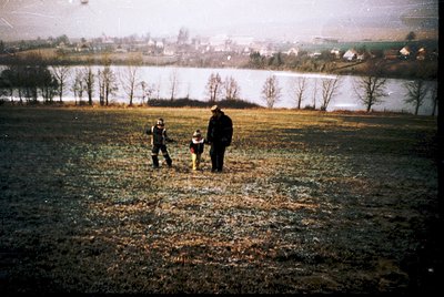 Vintage black-and-white photo of three figures walking along a snow-dusted field near a frozen lake, with rural houses in the...