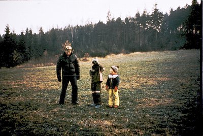 Family of three posing outdoors in a grassy field with forested background, likely mid-20th century. Adult wears vintage outd...