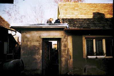 Vintage rural scene featuring a weathered concrete house with a sloped tiled roof. Two figures—likely men—stand on the roof, ...