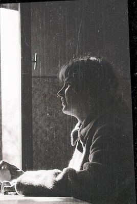 Black-and-white portrait of a person seated indoors, backlit by natural light, suggesting a mid-20th-century setting. The ind...
