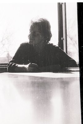 Portrait of an elderly woman seated indoors, leaning on a table near a window. She wears a dark, high-collared sweater and he...